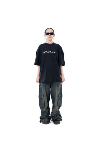A model stands against a solid white background wearing a black oversized t-shirt with a colorful graphic across the chest, paired with dark washed cargo pants and black shoes.
